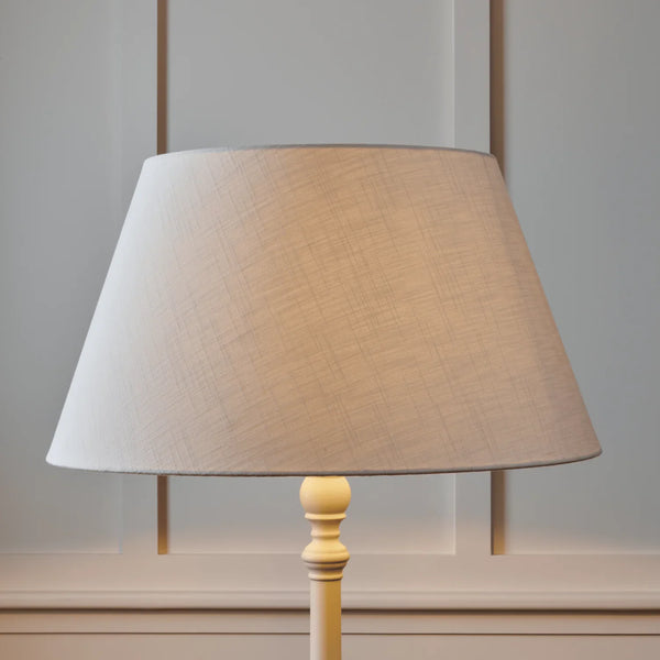 A table lamp with a tapered beige fabric shade and a light wood pedestal base, shown against a softly paneled gray wall.