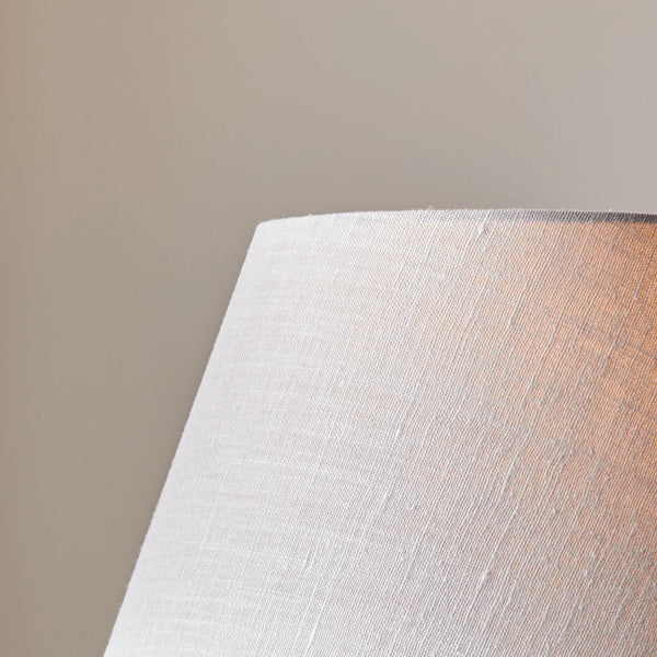 Close-up of a textured fabric lampshade in a warm off-white or light beige color, with a soft warm glow visible on the right side, set against a solid neutral beige background.