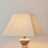 A table lamp with a large beige fabric drum shade and a distressed stone or wood base, shown against a textured beige wall.