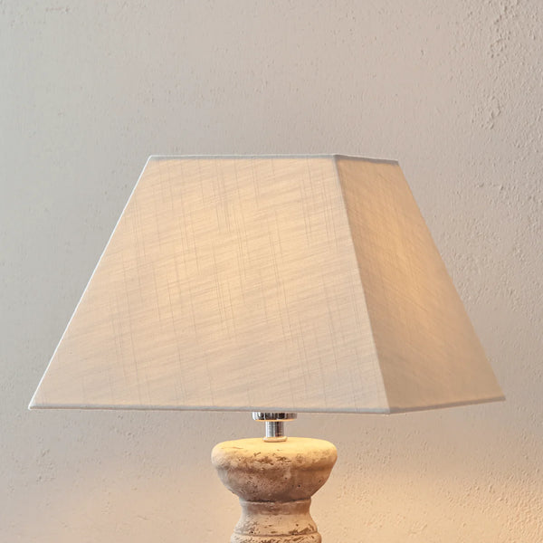 A table lamp with a large beige fabric drum shade and a distressed stone or wood base, shown against a textured beige wall.