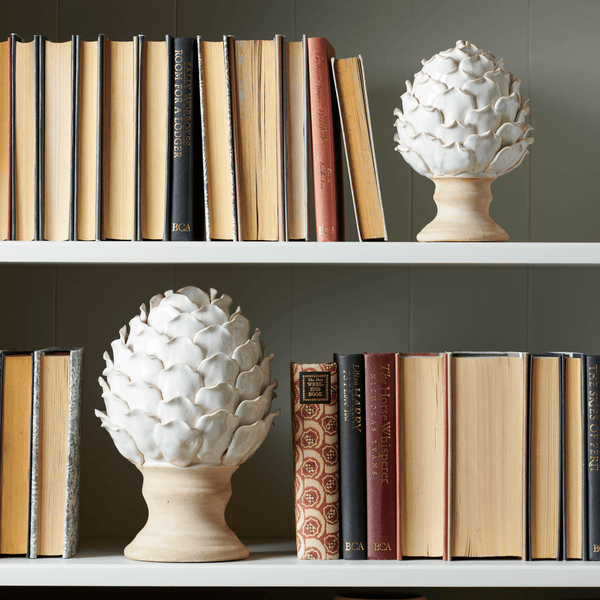 Two decorative white ceramic artichokes on beige pedestals, styled on white bookshelves next to vintage books against a dark gray paneled wall.