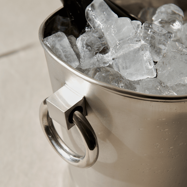 A polished silver ice bucket filled with clear ice, shown close up on a light beige surface.