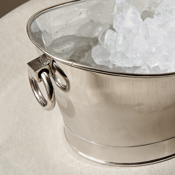A polished silver ice bucket filled with clear ice, shown close up on a light beige surface.