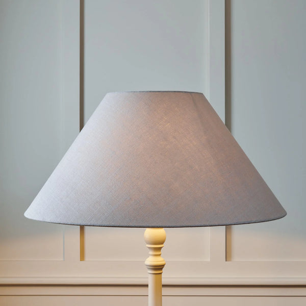 A table lamp with a large conical fabric shade in a light gray color and a light wood base, set against a pale gray paneled wall.