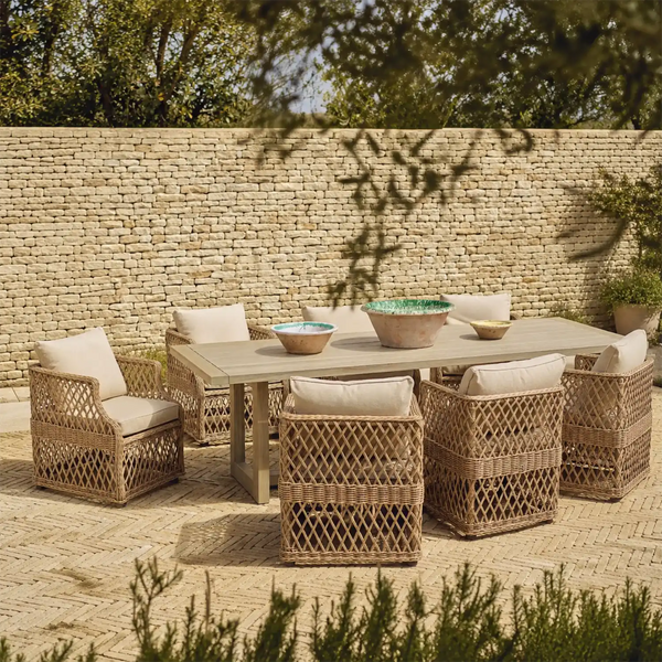 Outdoor dining set with a rectangular light wood table and five woven rattan armchairs with cream cushions, arranged on a paved patio in front of a stone wall.