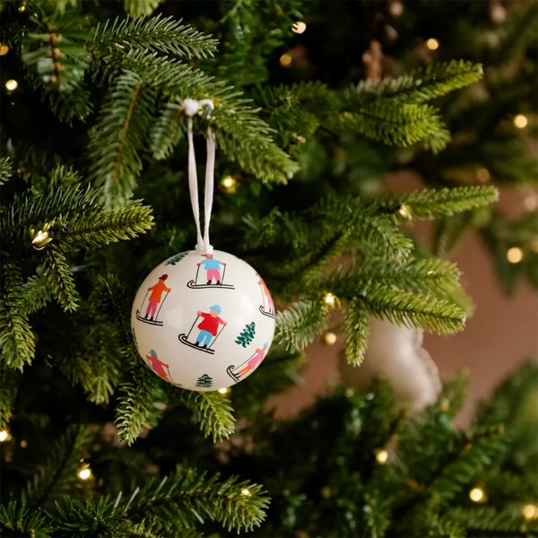 A round white Christmas ornament printed with colorful skiers, hanging from a green Christmas tree with warm string lights in the background.