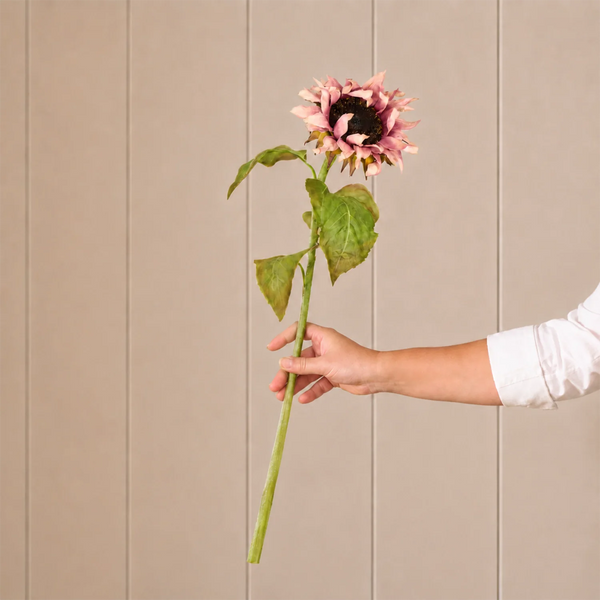 A single artificial-looking pink sunflower with a dark brown center, green stem, and leaves, held by a person's hand against a beige paneled background.
