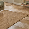 A natural jute area rug in a warm tan color, styled in a living room setting on a light wood herringbone floor next to a gray armchair and a wooden side table.