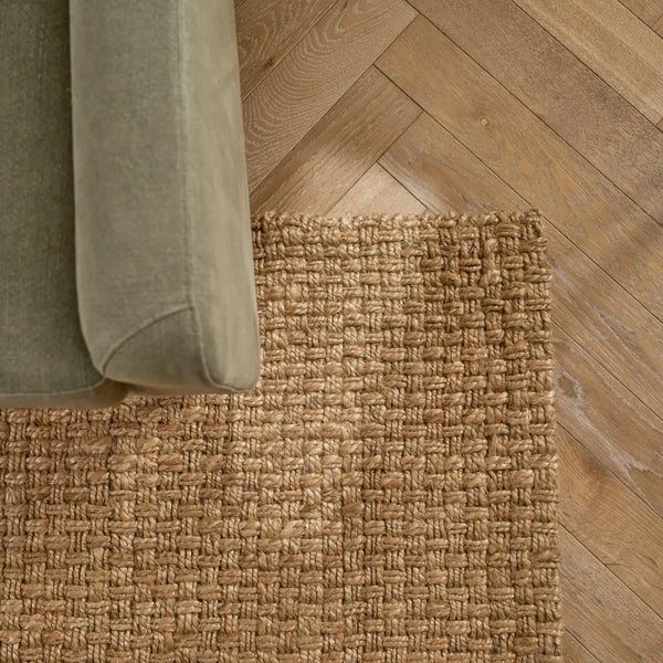 A close-up of a woven natural fiber area rug in a warm tan color, shown under the corner of a muted sage green upholstered chair on a light brown herringbone wood floor.