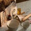 A styled baking or tea preparation scene on a marble countertop with a wicker basket of hay, a bag of organic chocolate chips, a small bottle of amber liquid, a jar of honey, a wooden rolling pin, a whisk, and two ceramic bowls.