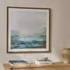 A framed abstract landscape painting with blue, green, and gray tones, displayed on a beige paneled wall above a wooden console table with books and a ceramic vase.