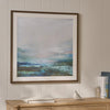 A framed abstract landscape art print with a brown frame and white mat, hanging on a beige wood-paneled wall above a light wood console table with books and a small bird figurine.