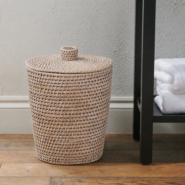 A natural woven rattan bin with a matching lid and top loop, set on a wooden floor next to a black shelving unit with white towels.